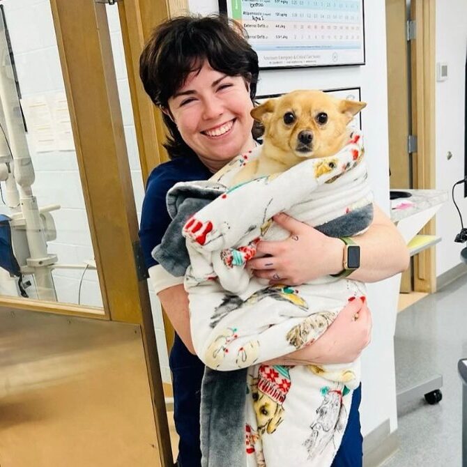 A smiling woman in scrubs holds a small tan dog wrapped in a fleece blanket inside a veterinary clinic, standing by a door with medical equipment visible in the background.