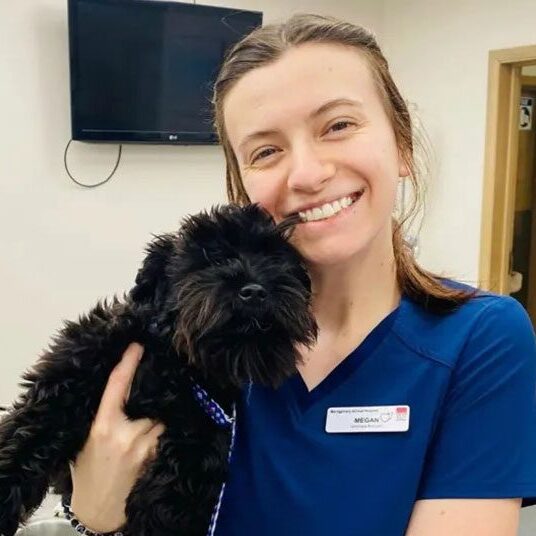 The veterinarian smiles warmly, cradling a small black dog in an office setting. They're wearing a name tag and a dark blue shirt, radiating care and professionalism.