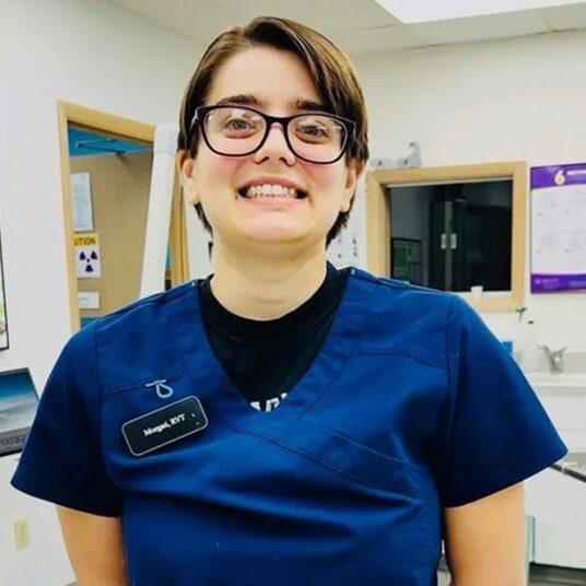 A veterinarian wearing glasses and a blue scrub top stands smiling in a veterinary clinic. The environment includes medical equipment, a laptop, and informational posters on the walls.