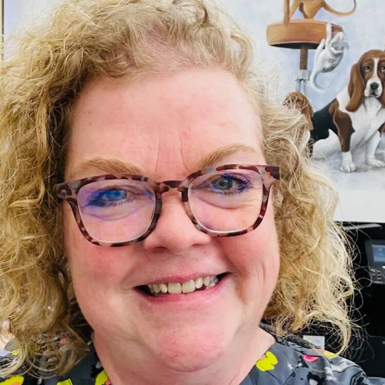A person with curly blonde hair and glasses, likely a veterinarian, smiles at the camera. In the background, a mural depicts various animals, including a dog and a cat.