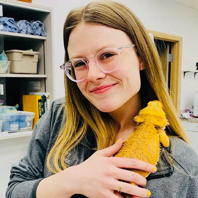 A smiling veterinarian with glasses holds a yellow bearded dragon lizard in a room filled with shelves and equipment.